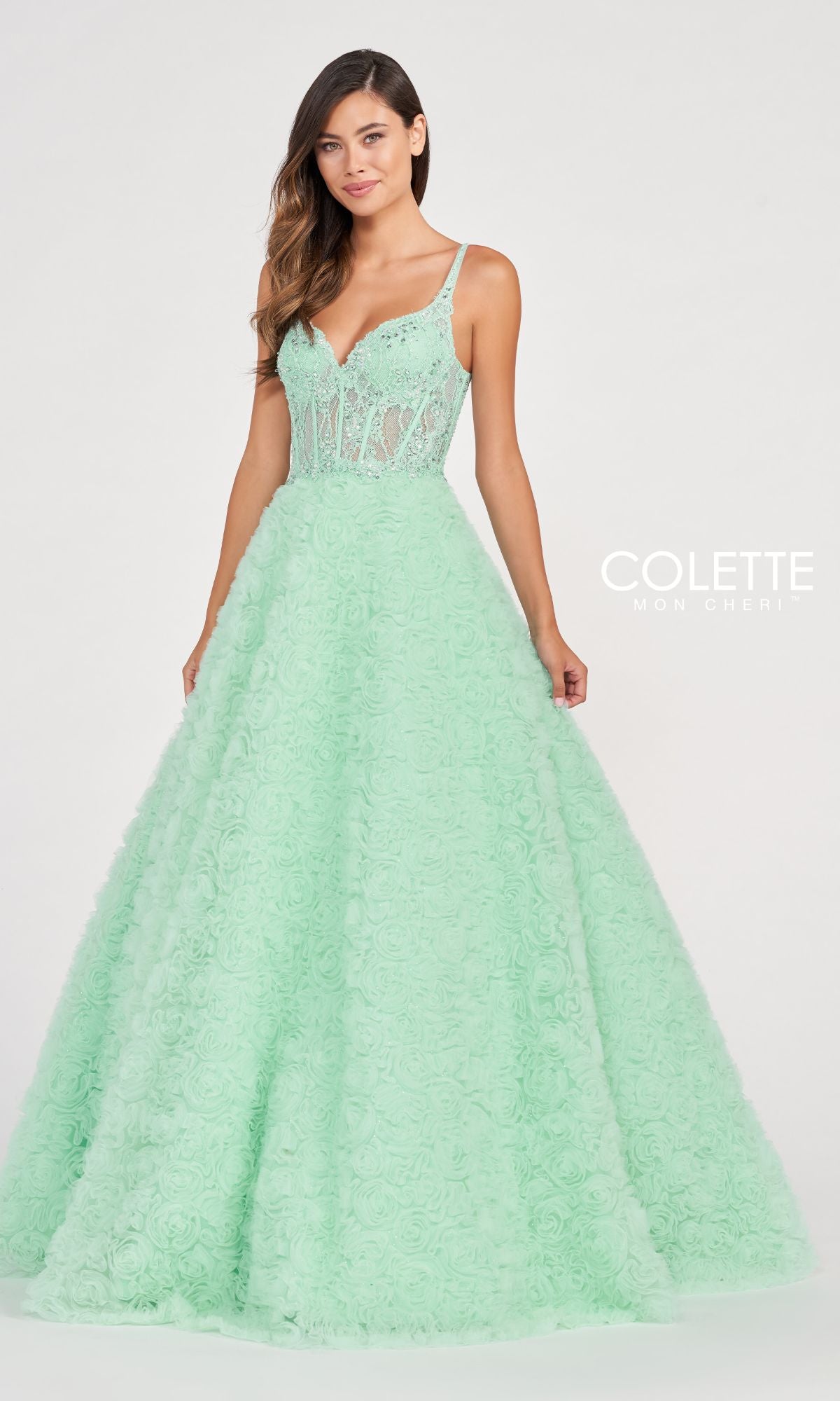 A woman wears the Colette Sheer-Bodice Rosette Beaded Lace Ball Gown, featuring a mint green hue, fitted sheer bodice, and floral textured skirt, as she stands against a plain light background.